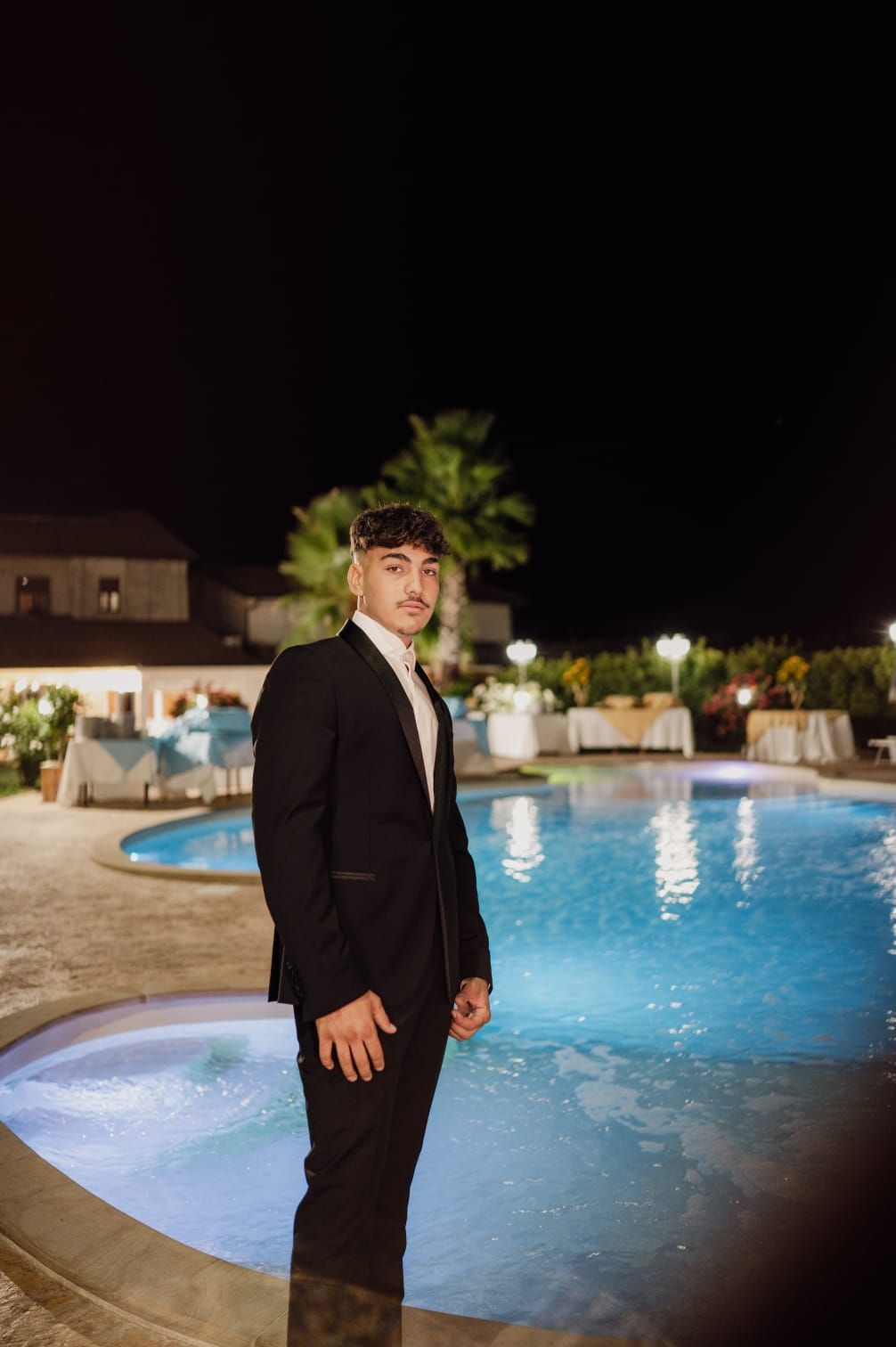 Un uomo in giacca e cravatta è in piedi vicino a una piscina di notte; sullo sfondo si vedono edifici e palme.