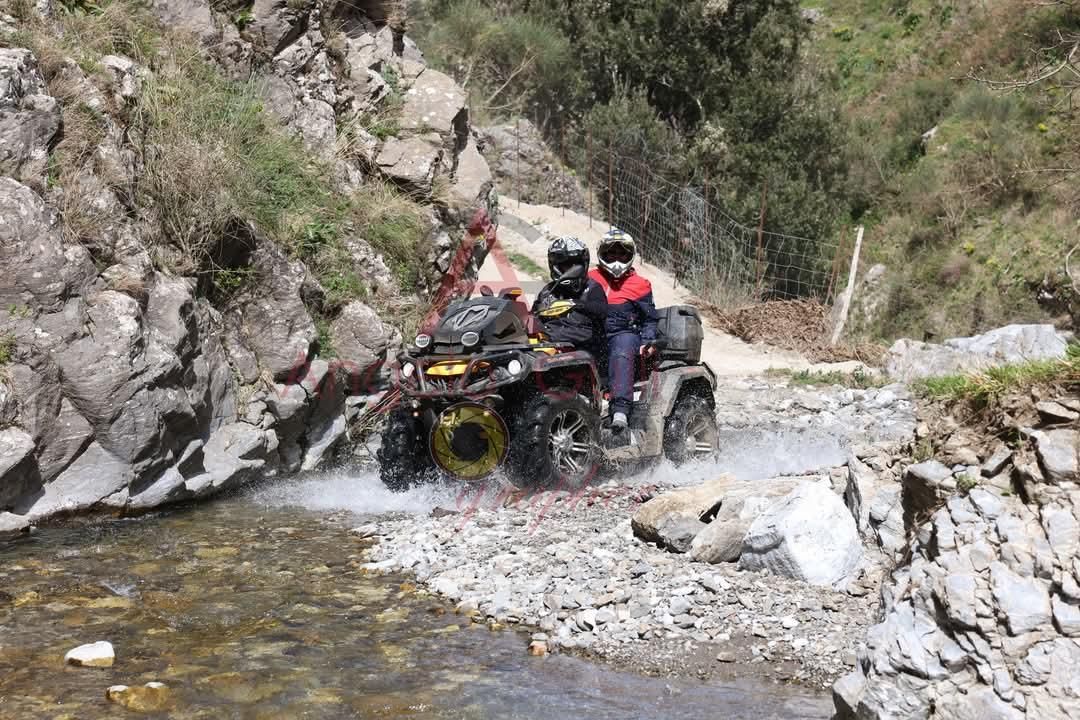 Un quad attraversa un fiume roccioso e poco profondo. Due persone con il casco viaggiano sul quad; si vede un pendio roccioso.
