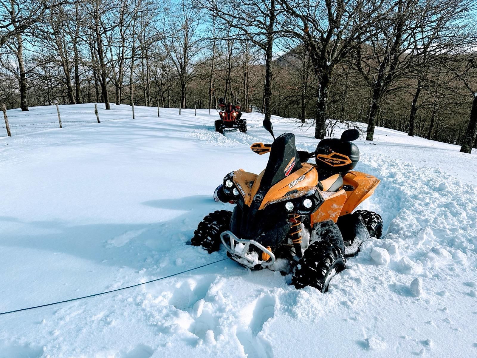 Un quad arancione bloccato nella neve alta, un altro quad in lontananza, un campo innevato con alberi.