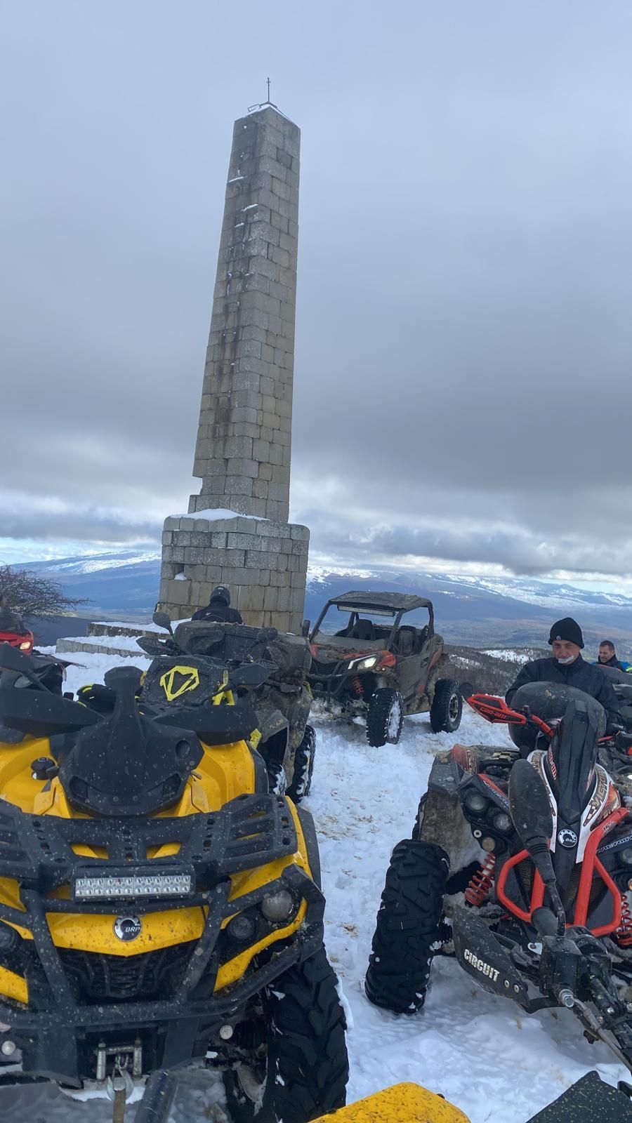 Quad vicino a un monumento in pietra sulla cima di una montagna innevata. Piloti in abiti scuri, cielo coperto.