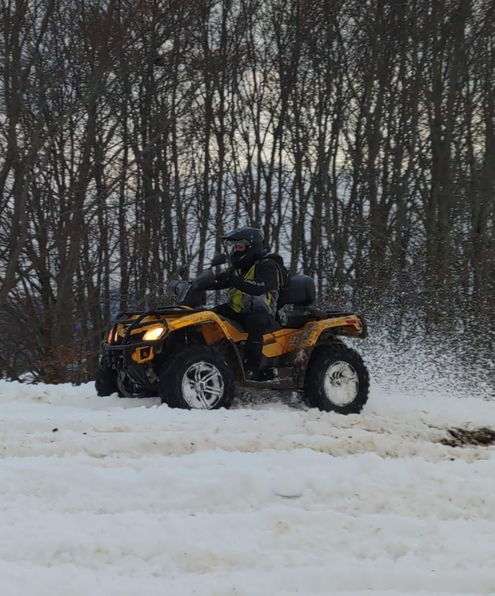 Un quad giallo che guida sulla neve, una persona con il casco. Alberi sullo sfondo.