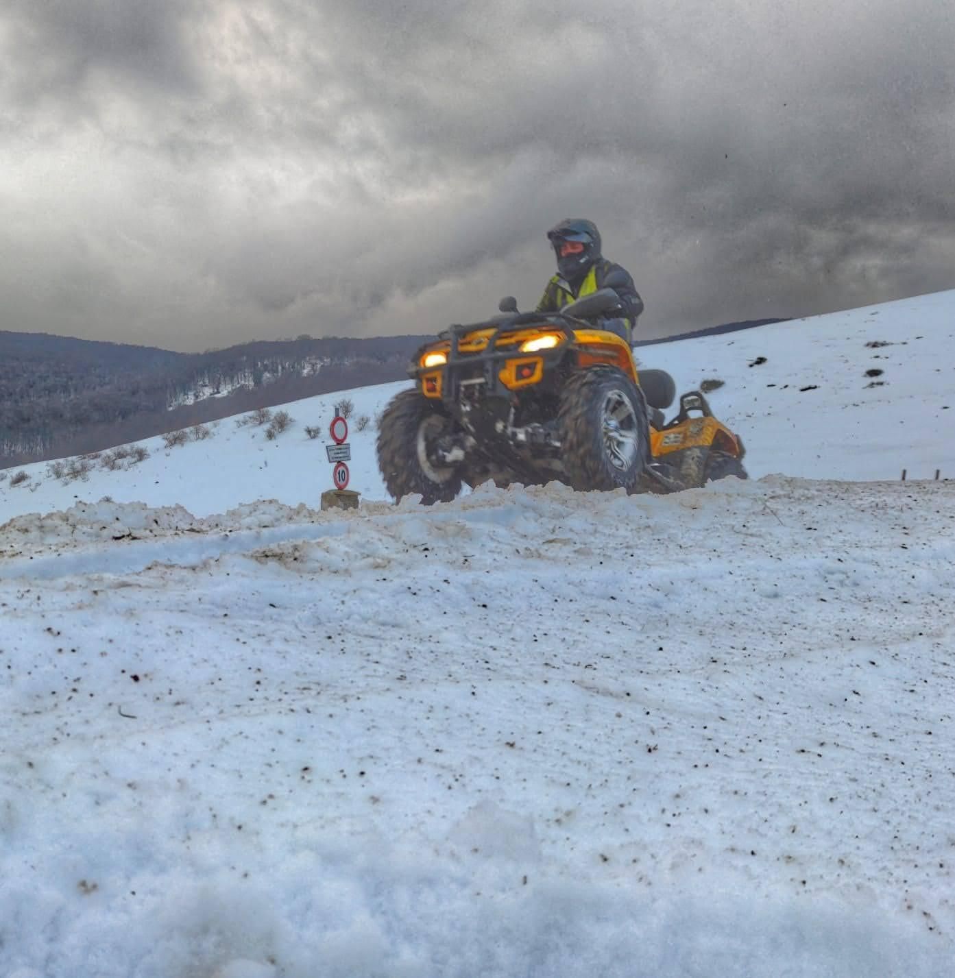 Persona alla guida di un quad giallo su un pendio innevato sotto un cielo nuvoloso.