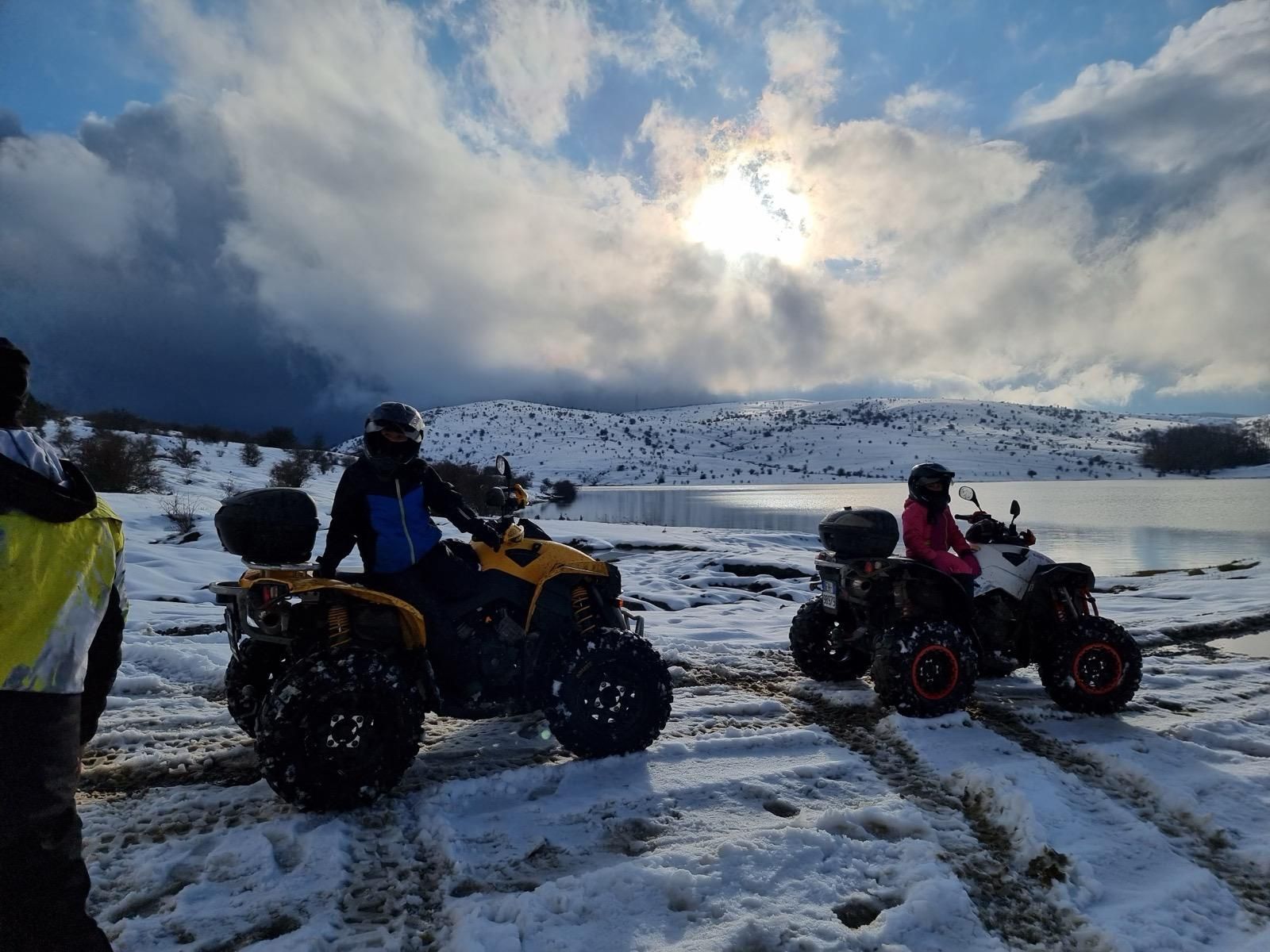 Due persone su quad in un paesaggio innevato vicino a un lago, sotto un cielo nuvoloso.