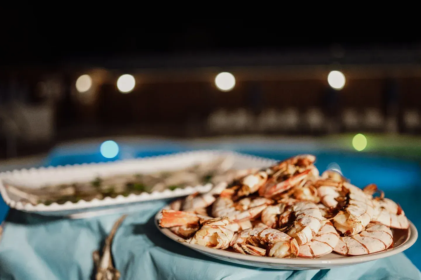 Piatti di gamberi cotti, vicino a una piscina, di notte. Luci bokeh sullo sfondo.