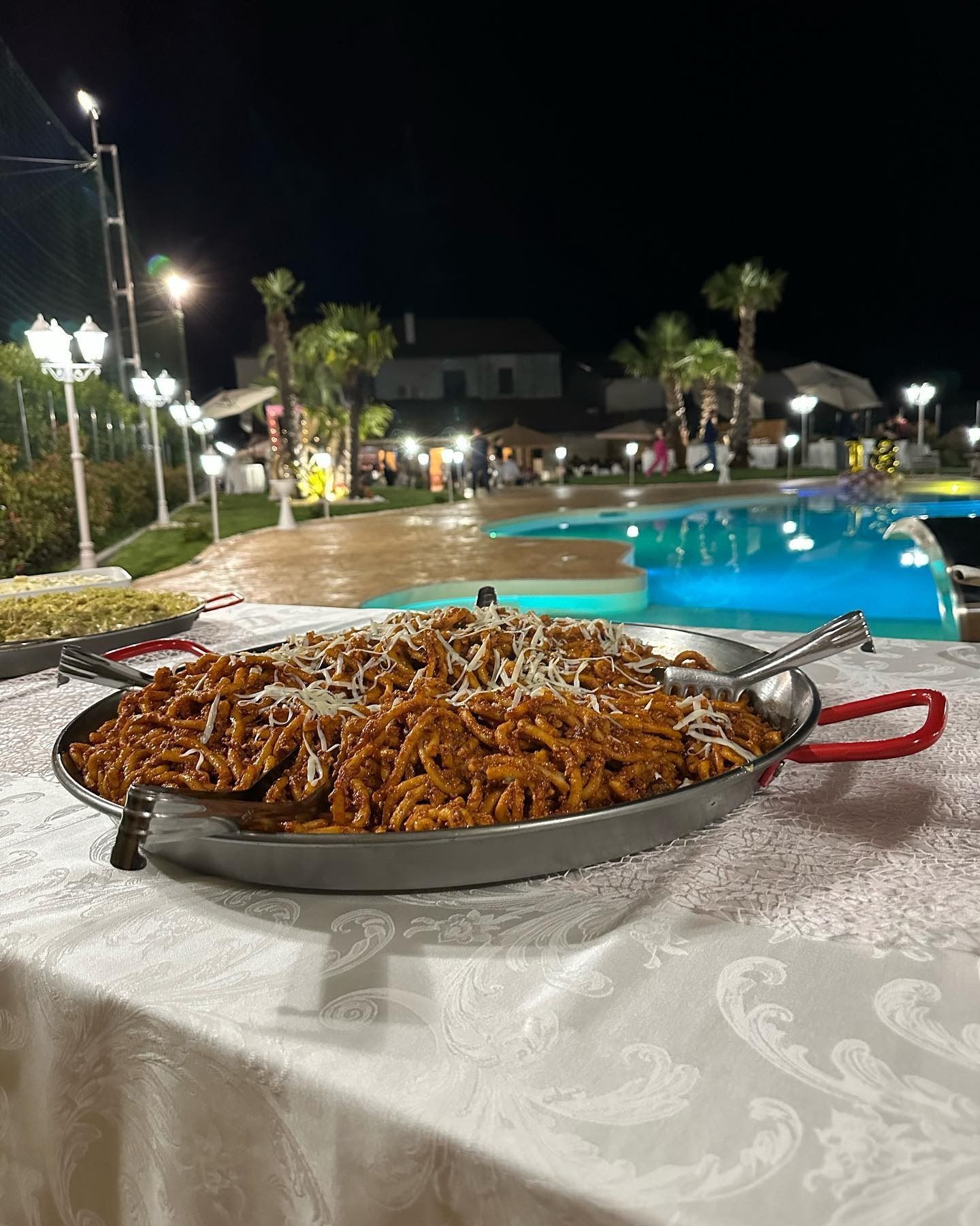 Un grande piatto di pasta sul tavolo, a bordo piscina di sera. Il cibo è marrone, condito con formaggio.