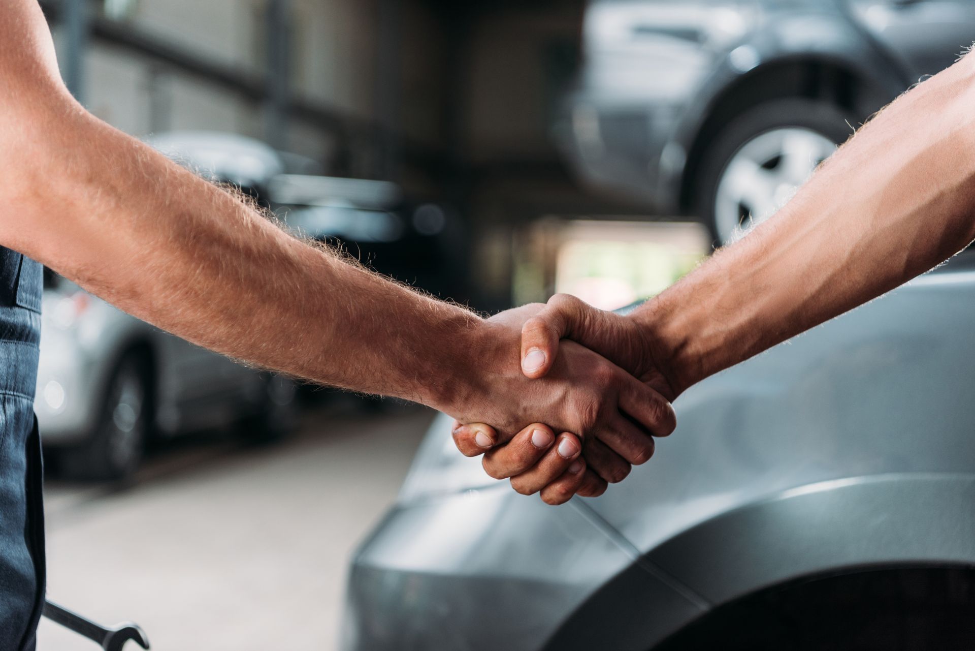 Two people shaking hands in an auto repair shop with cars visible in the background.
