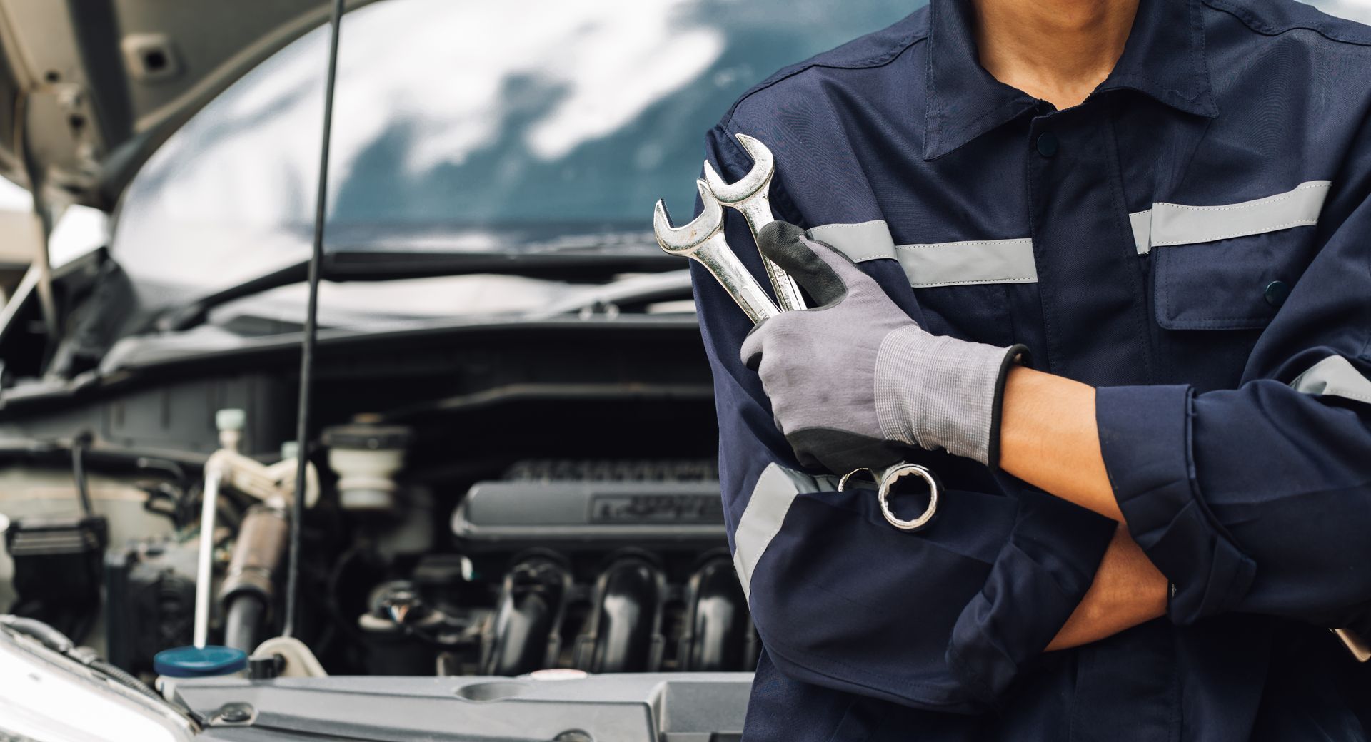 Mechanic holding wrenches in front of an open car engine hood.