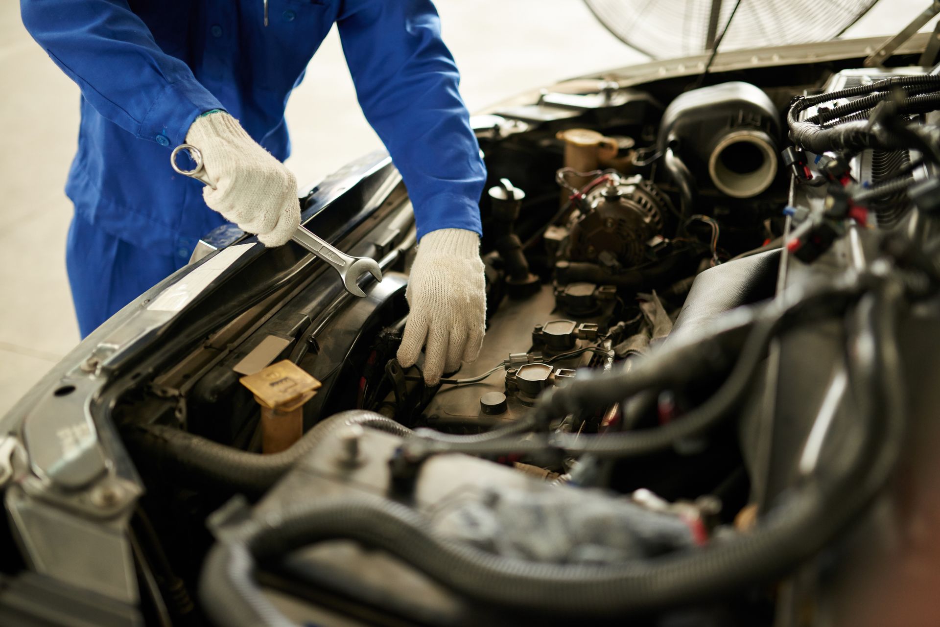 Mechanic in blue jumpsuit works on car engine with a wrench. White gloves are worn.