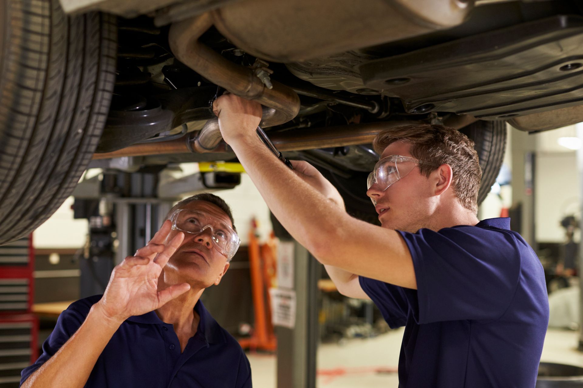 Mechanics repairing car exhaust system under lift in professional auto repair garage setting.