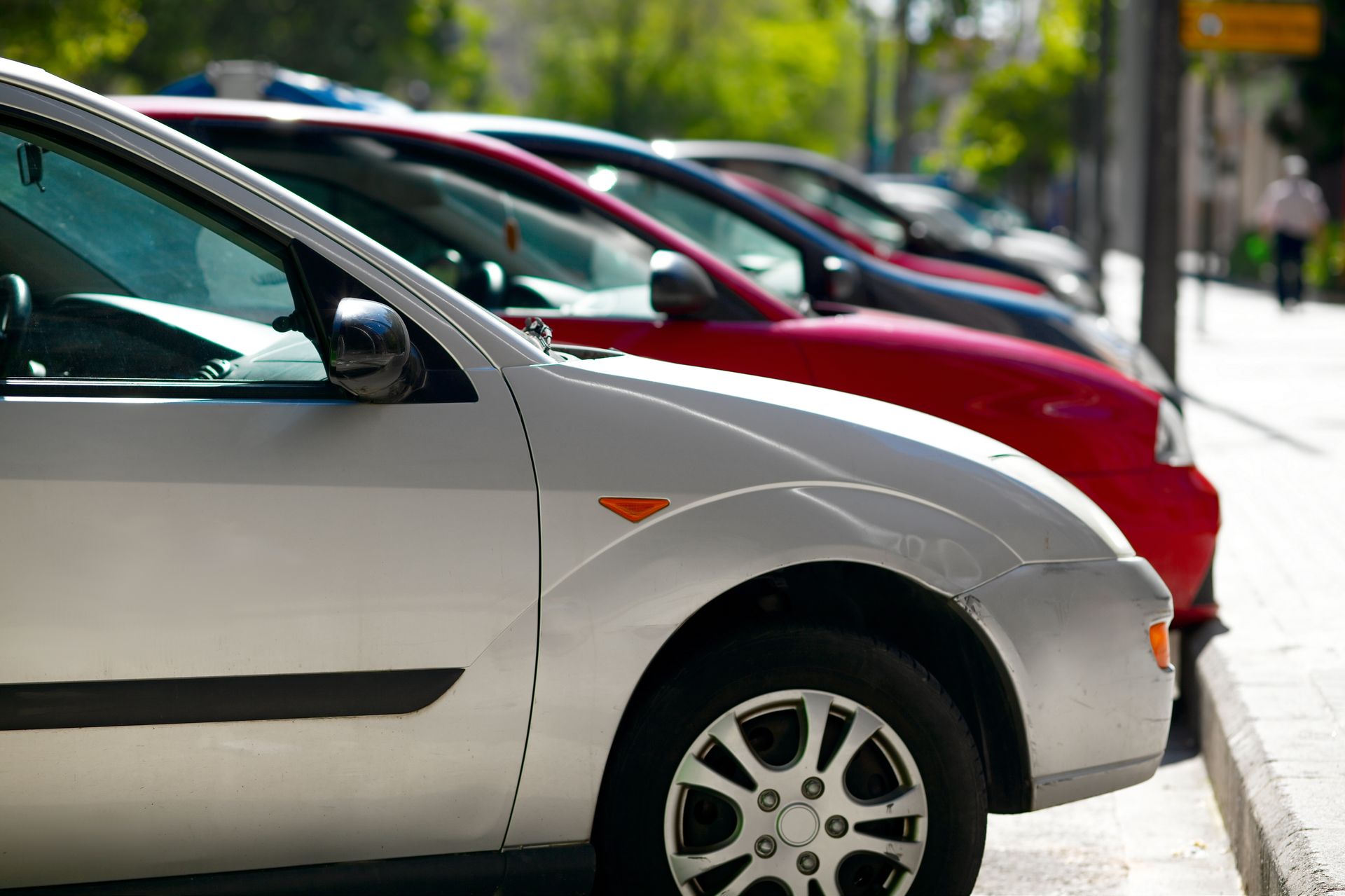 A line of cars parked along a city curb, with a silver car in the immediate foreground and a red car behind it.