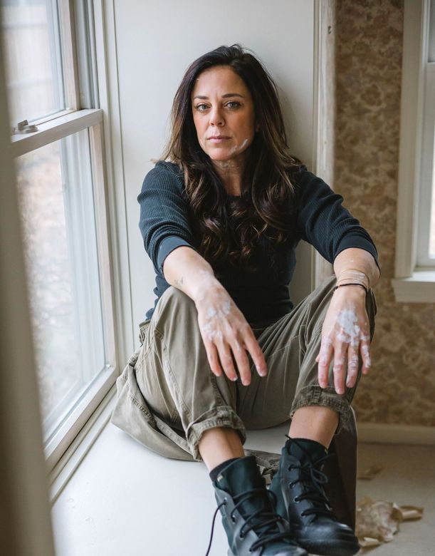 A woman in overalls and gloves is sitting on the floor in front of a wall.