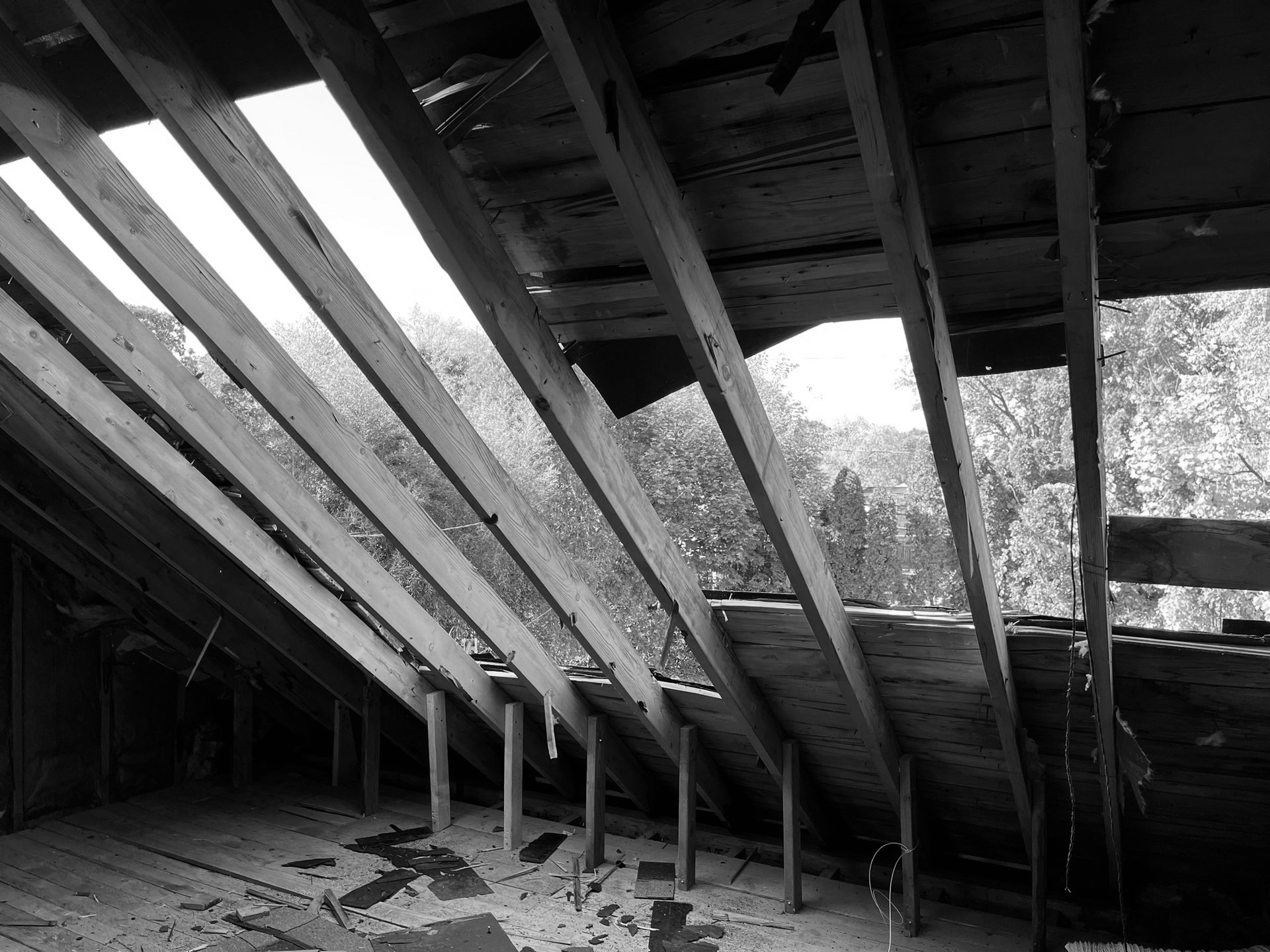 An attic interior with exposed wooden rafters and large sections of the roof missing, revealing trees in the background.