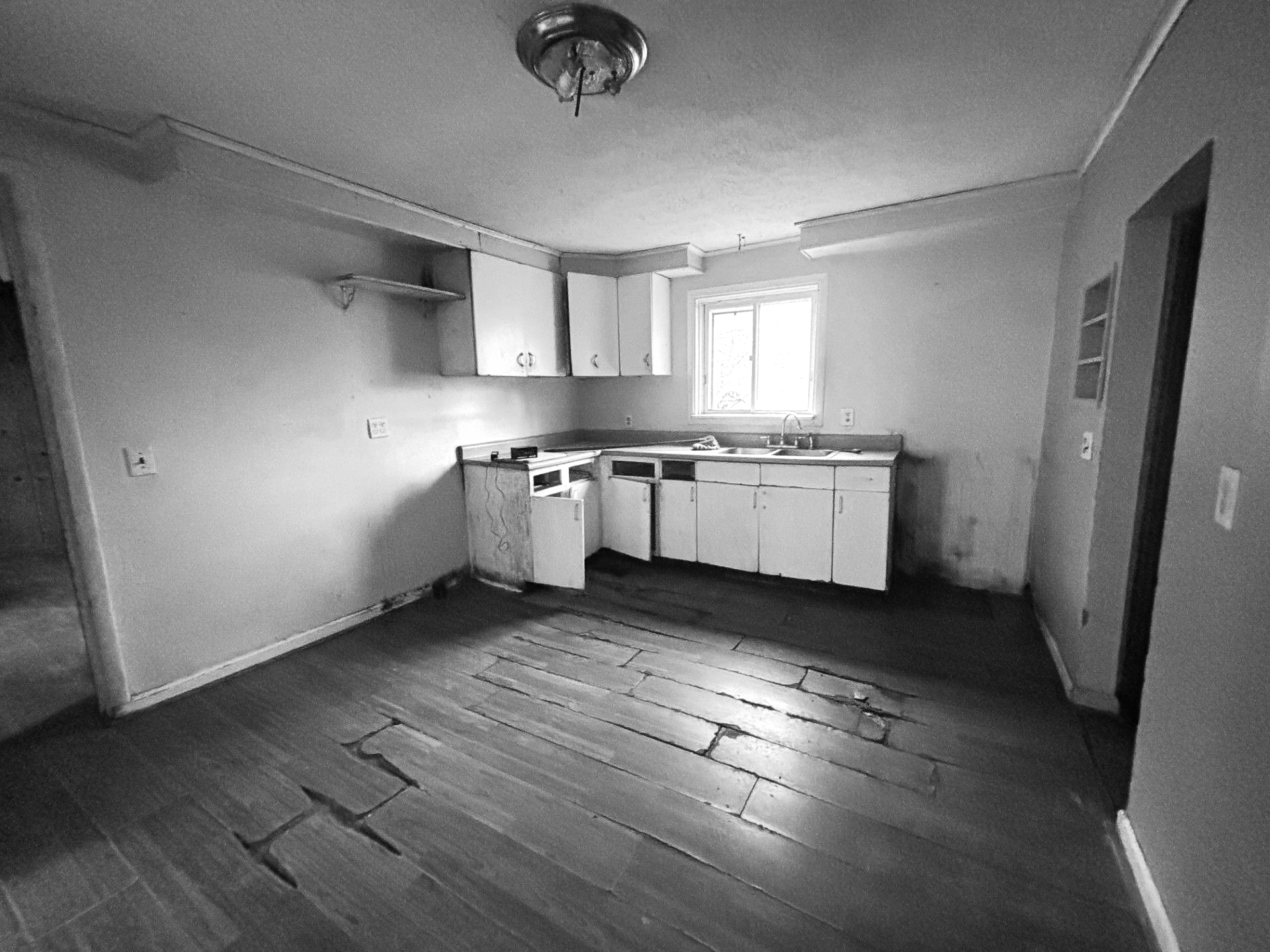 A monochromatic, high-angle view of a kitchen in disrepair, featuring bare walls, wood-plank flooring, and white cabinets.