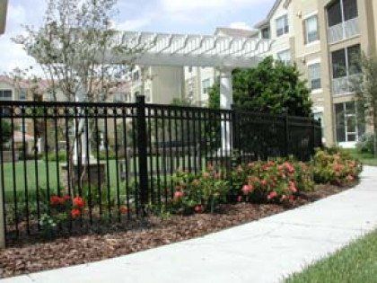 A black fence is surrounded by flowers and a sidewalk in front of a building.