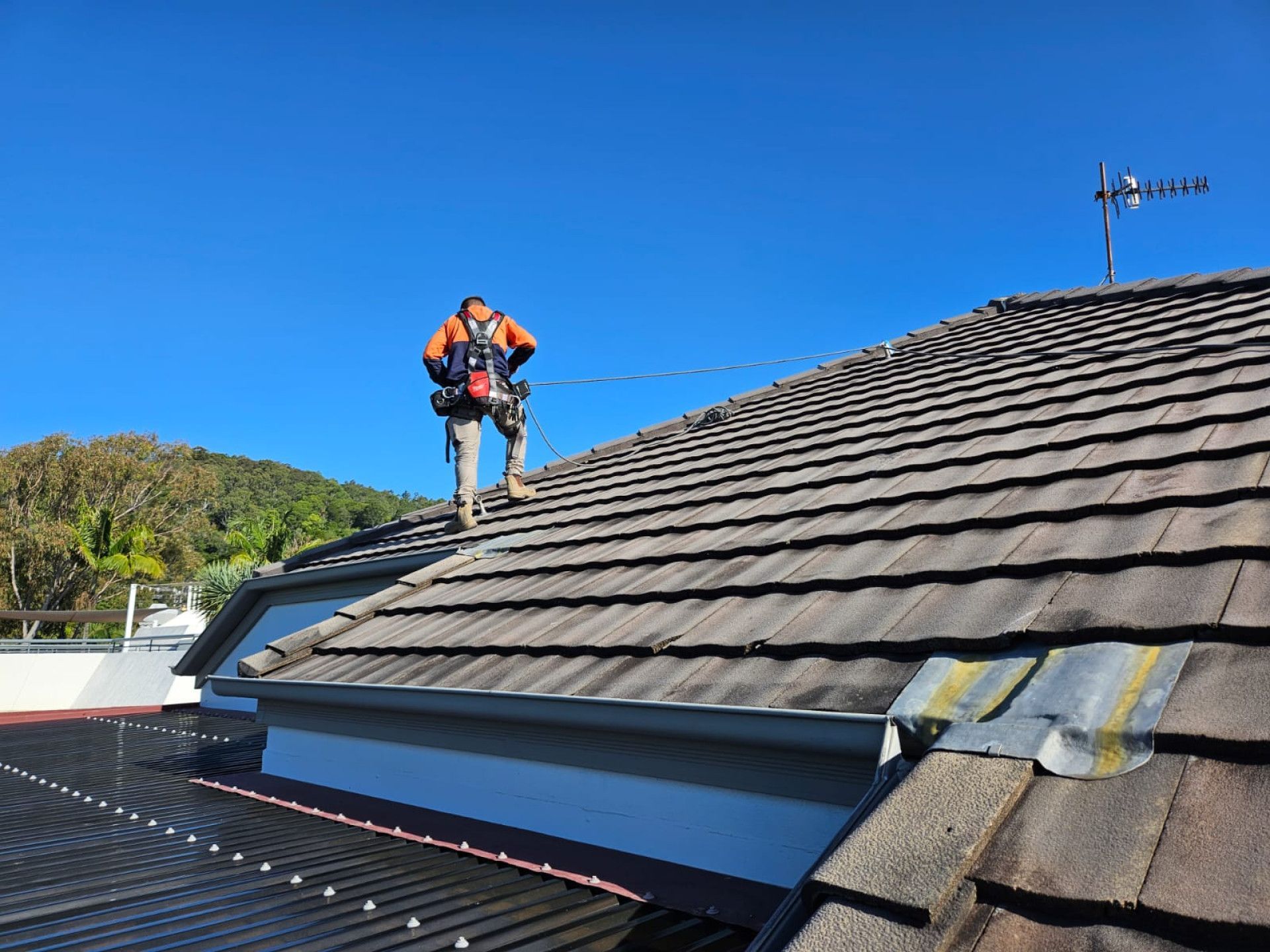 Person in safety gear on a tiled roof under a blue sky, performing roofing work. — Australian Asset Compliance in Glanmire, QLD