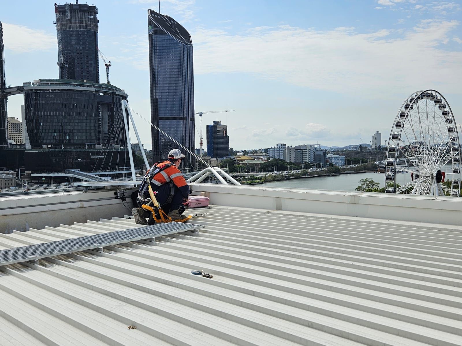 Roofer in safety gear installing metal on a white corrugated roof with a city skyline and Ferris wheel in the background. — Australian Asset Compliance in Glanmire, QLD