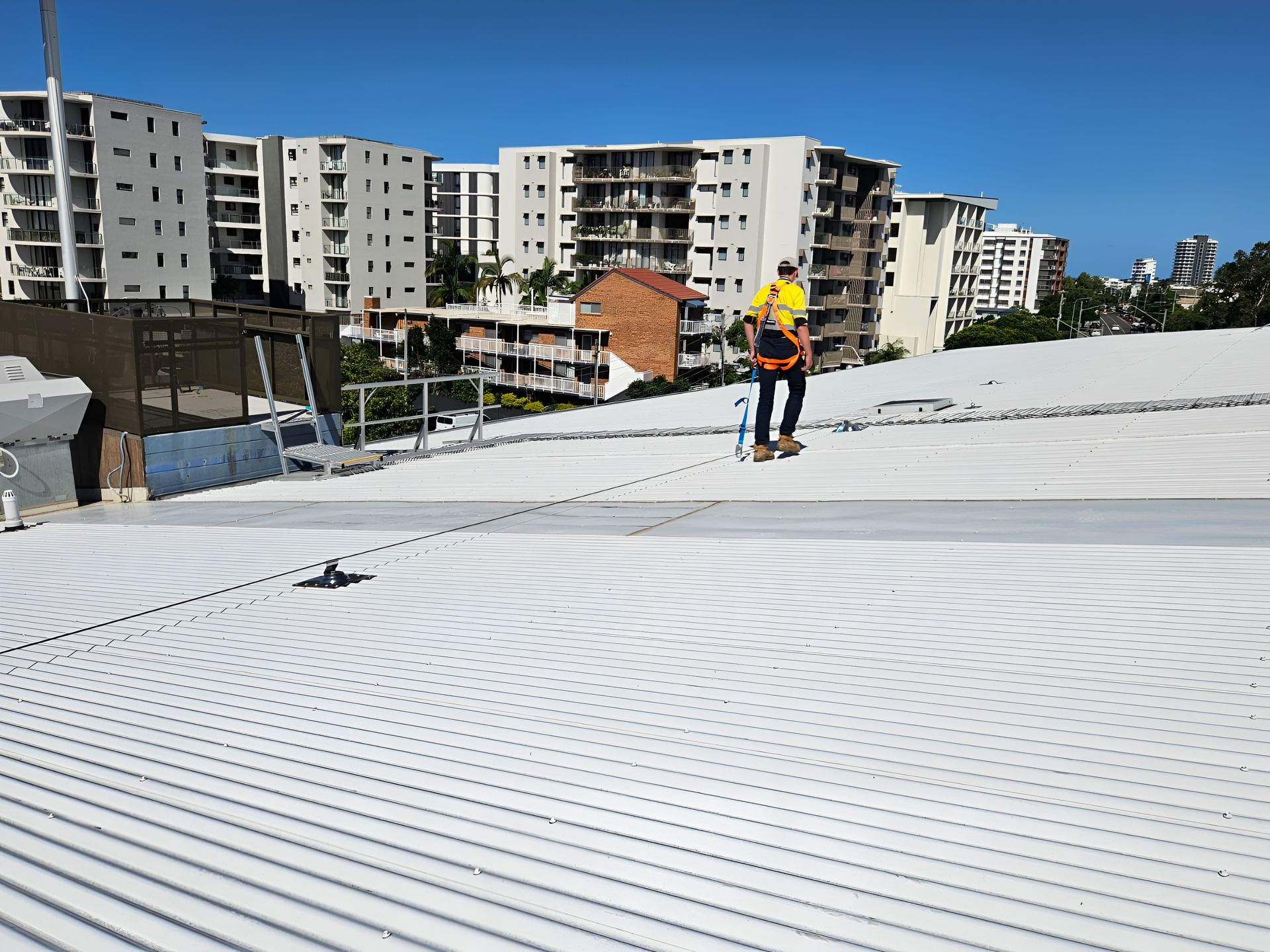 Roofer in safety gear installing metal on a white corrugated roof with a city skyline in the background. — Australian Asset Compliance in Glanmire, QLD