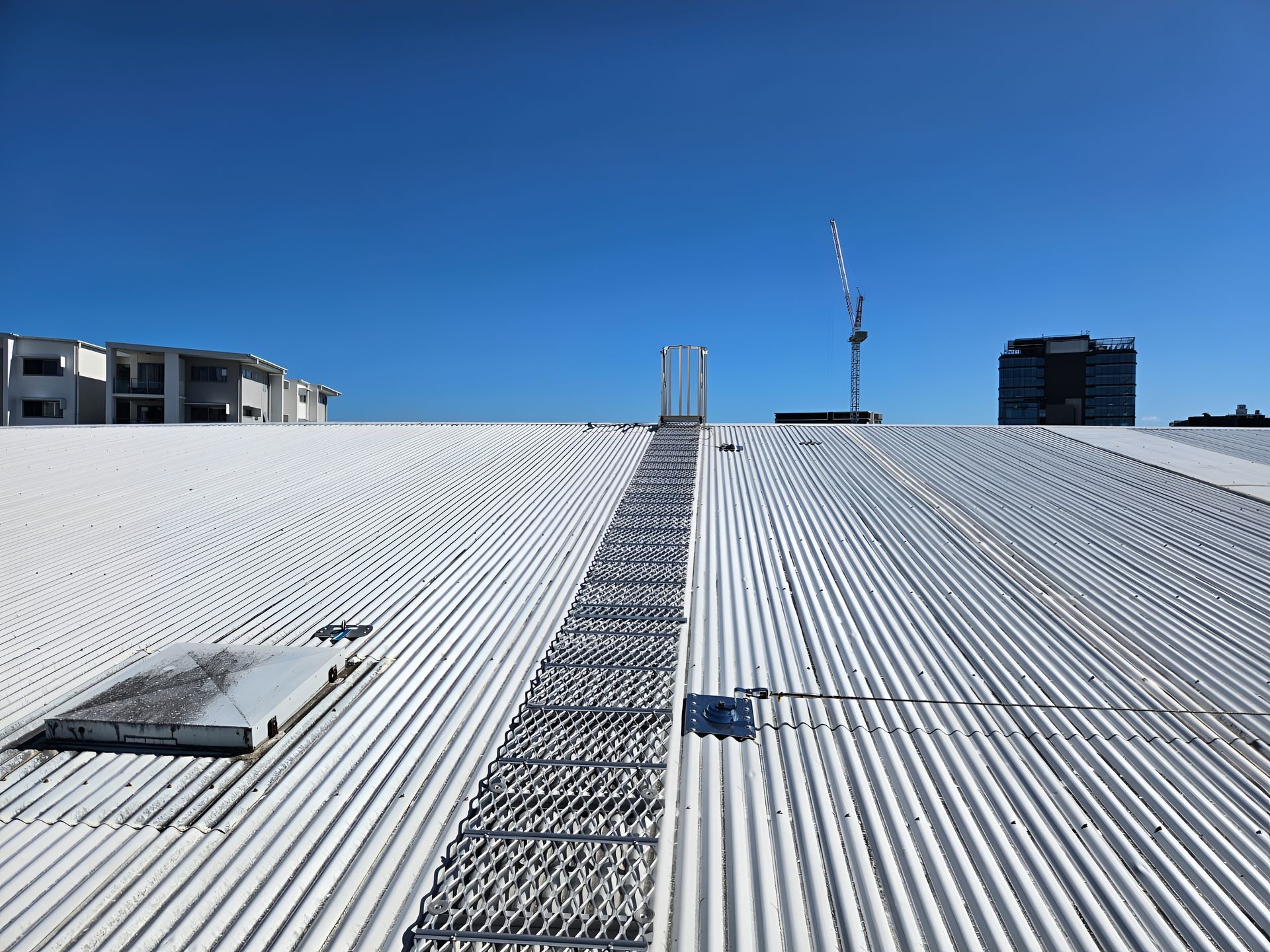 White corrugated roof with a metal ladder leading toward a crane and buildings under a blue sky. — Australian Asset Compliance in Brisbane, QLD