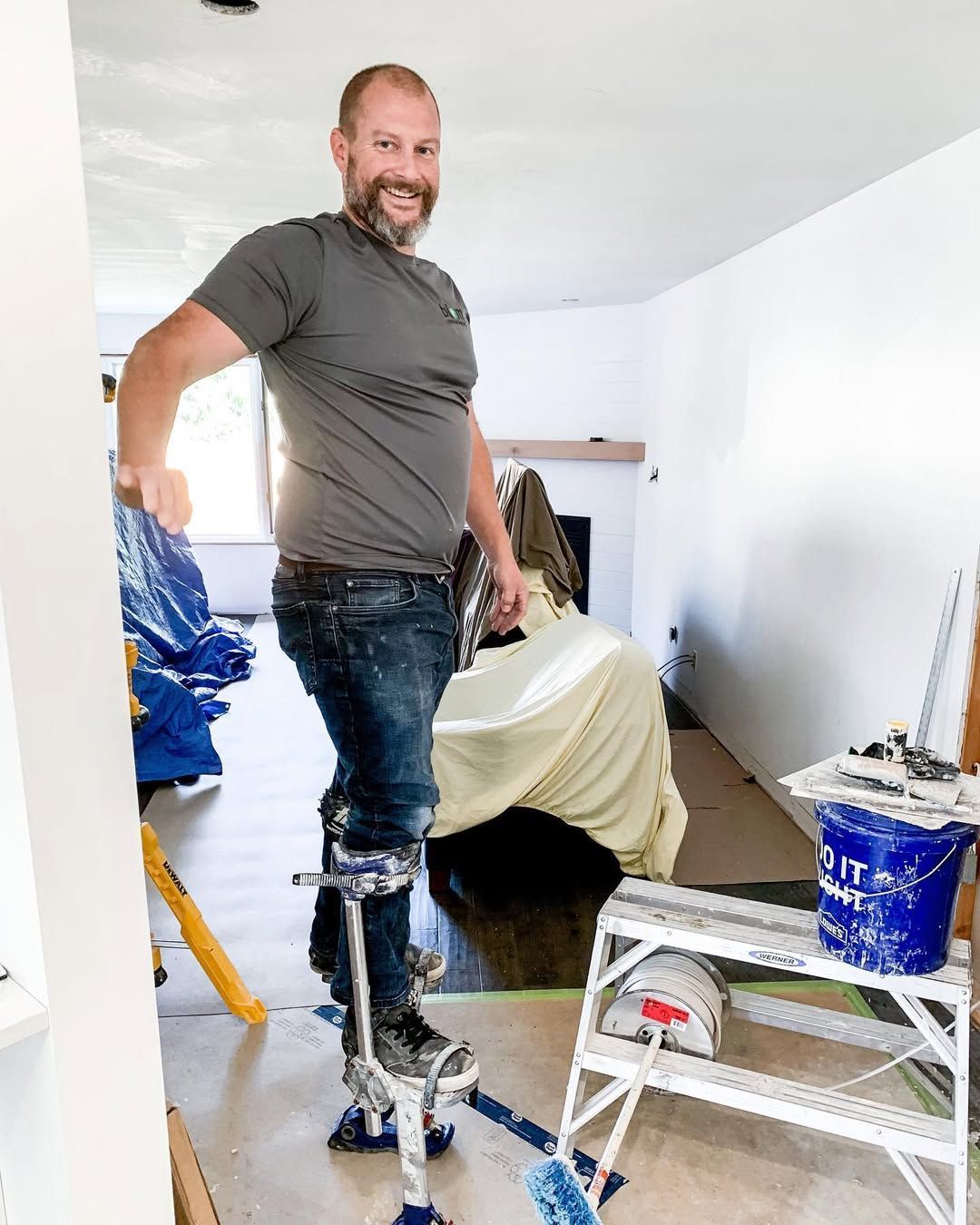 Man on stilts in room under construction smiles, leaning back. Tools and covered furniture are present.