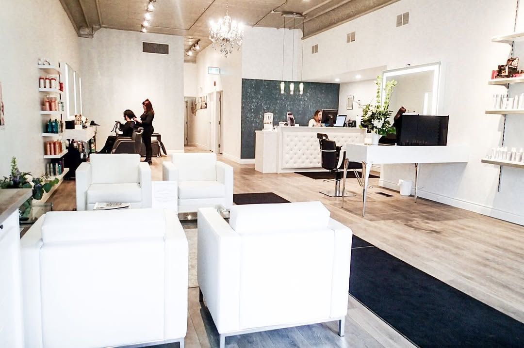 Hair salon interior with white furniture, stylists and clients, and a reception desk.