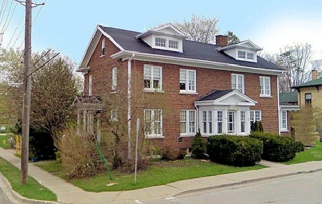 Brick two-story house with white trim, dormers, and bay window, on a corner lot with green lawn.