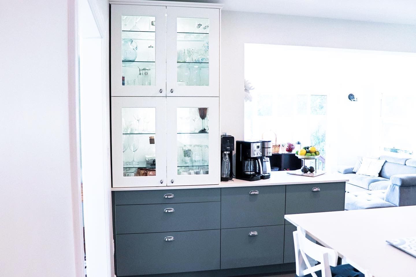 Kitchen cabinets with glass-front uppers, dark gray lowers, coffee maker on counter, bright window in background.