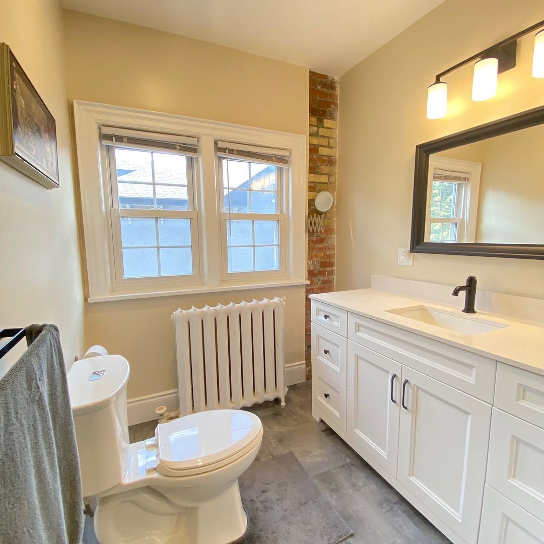 Bathroom with white vanity, toilet, window, brick wall, and gray floor.