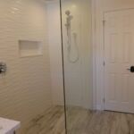 A modern bathroom with a glass shower, white tiled walls, and a wood-look floor.