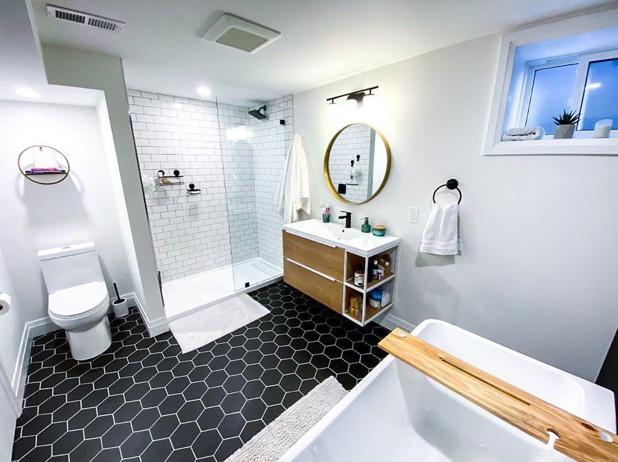 Modern bathroom: black hexagon tile floor, white shower, wood vanity, round mirror, and a soaking tub.