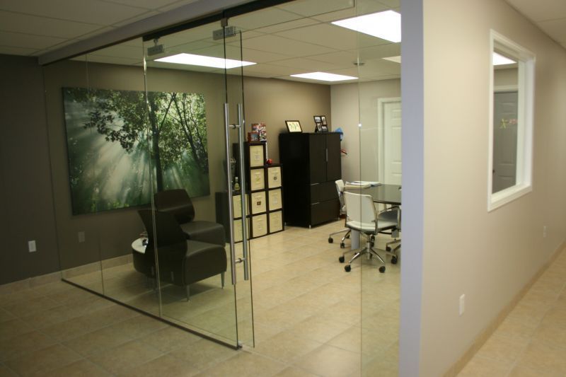 Glass-walled office with a waiting area, desk, art, and meeting area. Beige walls, a green tree art piece.