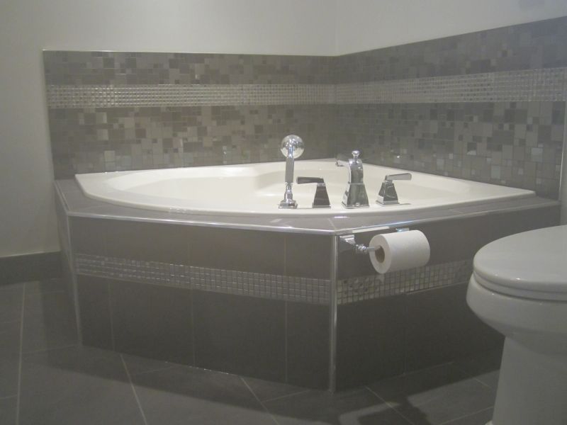 Corner bathtub with gray tile surround, mosaic accents, and chrome fixtures. Toilet is in the right.