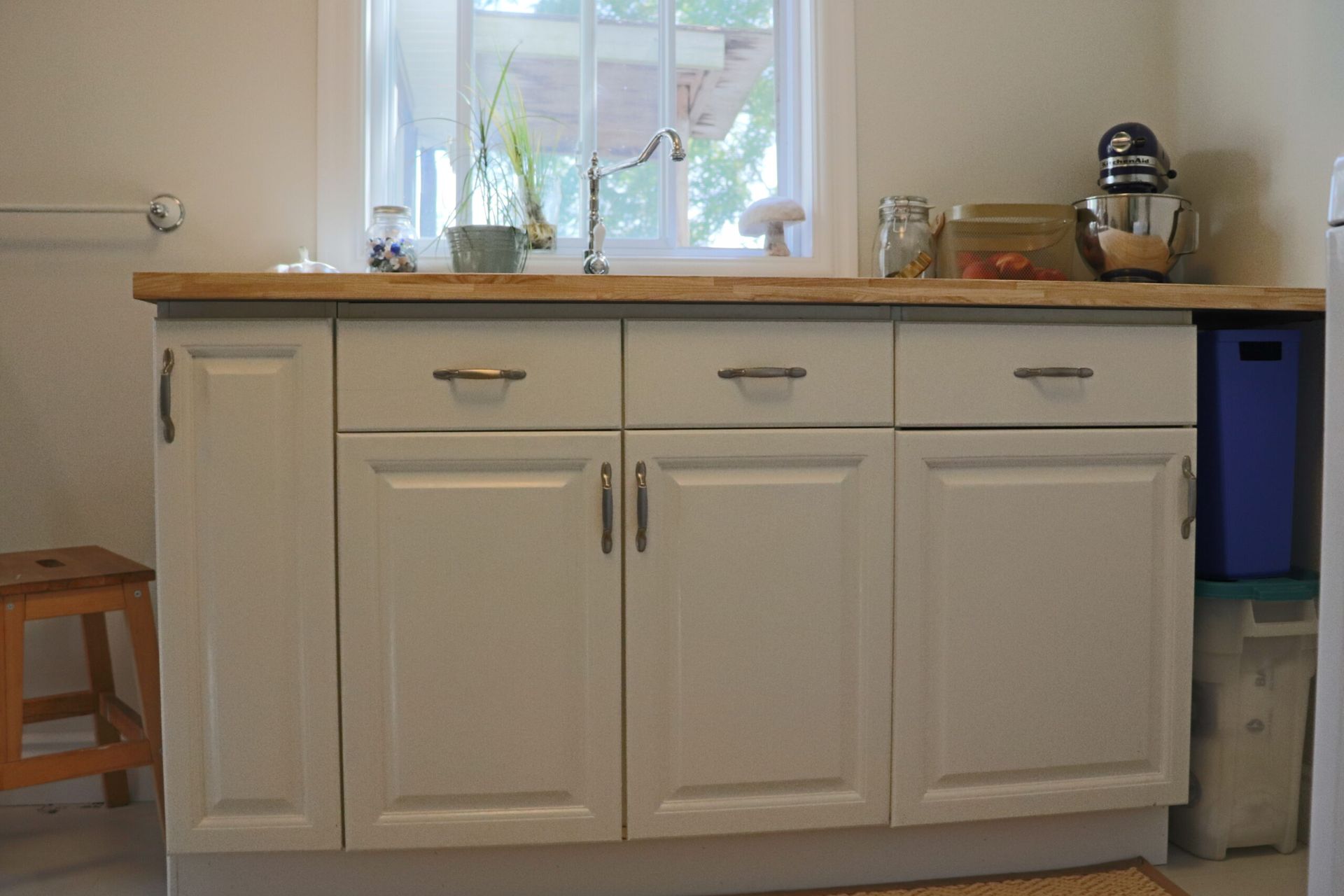 White kitchen cabinets with a wooden countertop under a window, including a blue bin on the right.
