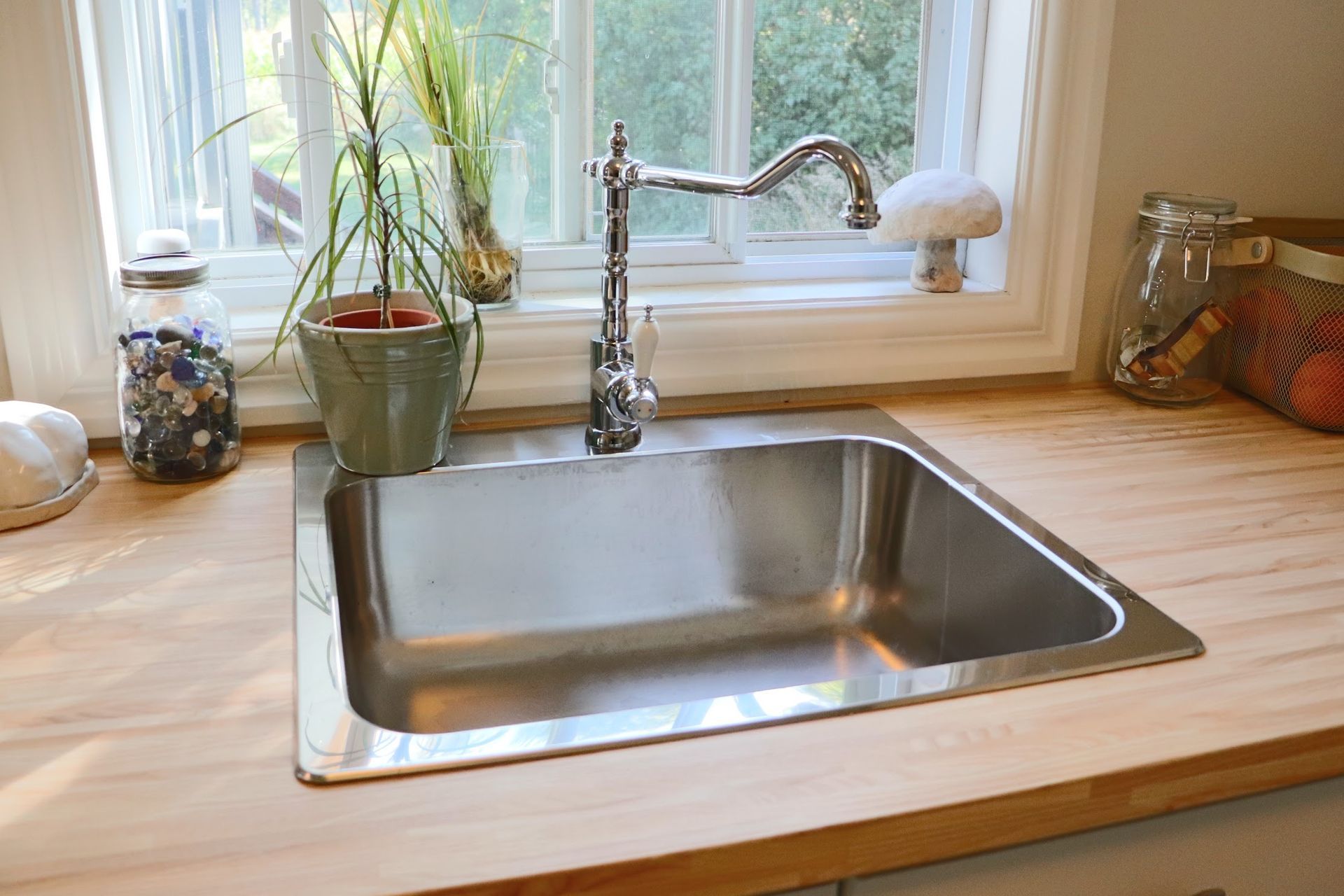 Stainless steel kitchen sink in front of a window, with a faucet, plants, and wooden countertop.