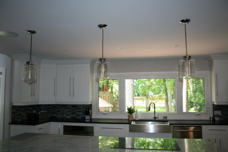 Kitchen with white cabinets, three pendant lights, stainless steel sink, and large windows overlooking trees.