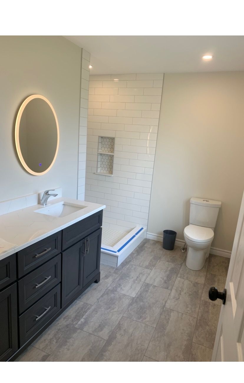 Modern bathroom: gray vanity, white countertop, oval mirror, glass shower, white subway tile, toilet, gray wood-look floor.