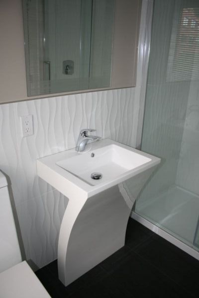 White pedestal sink in a modern bathroom with wave-patterned wall, mirror, and shower.