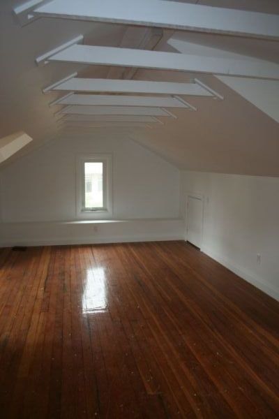 Bright, empty attic room with wood floors, white walls, and a small window; beams span the ceiling.