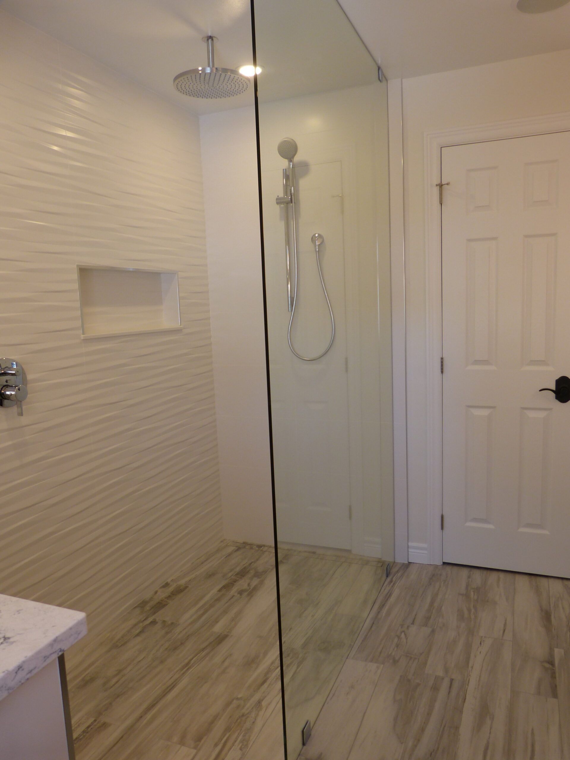 Modern white bathroom with textured wall, glass shower, wood-look floor. Door on the right.
