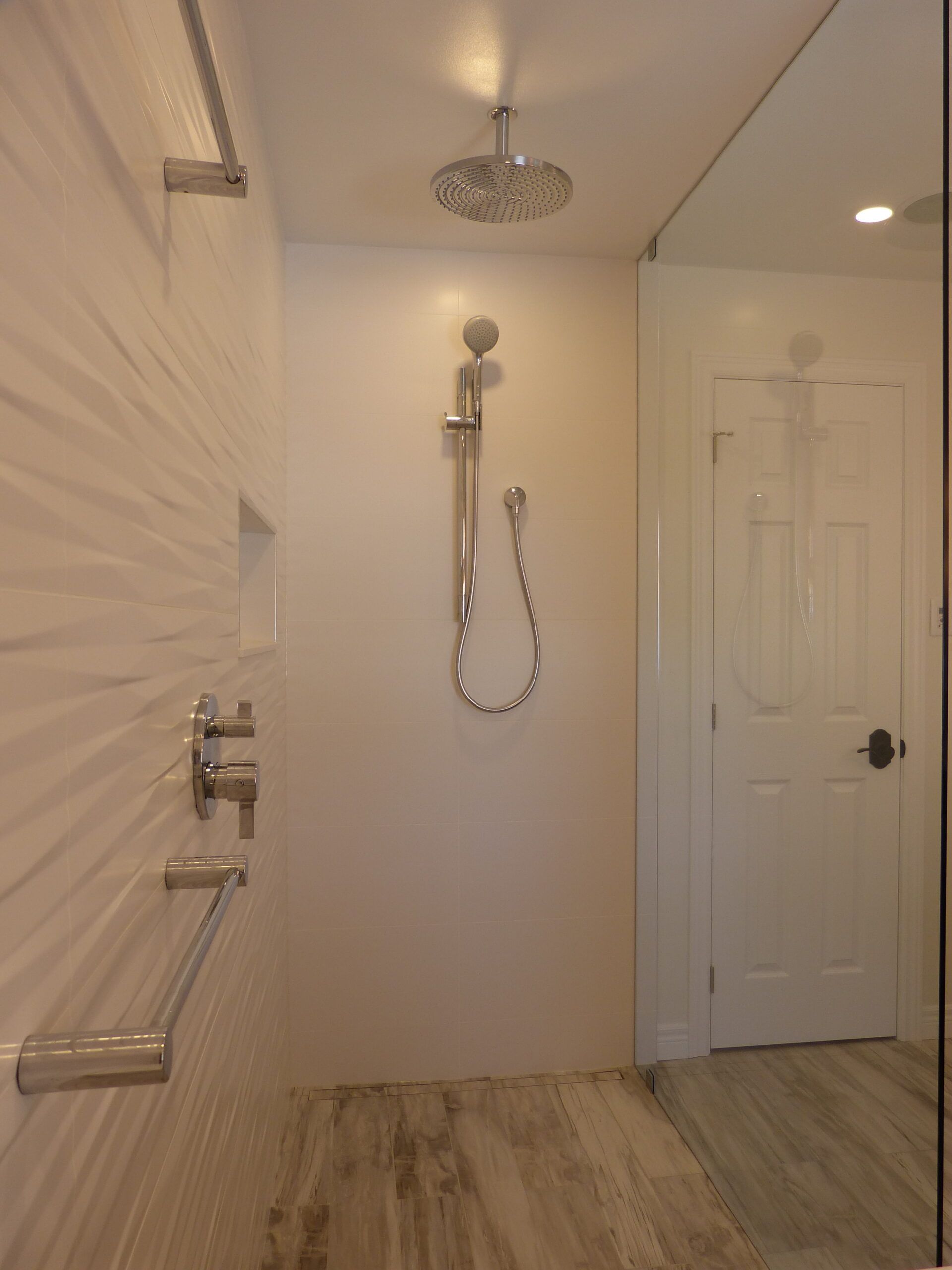 Modern white tiled shower with chrome fixtures, glass door, and a white door in the background.