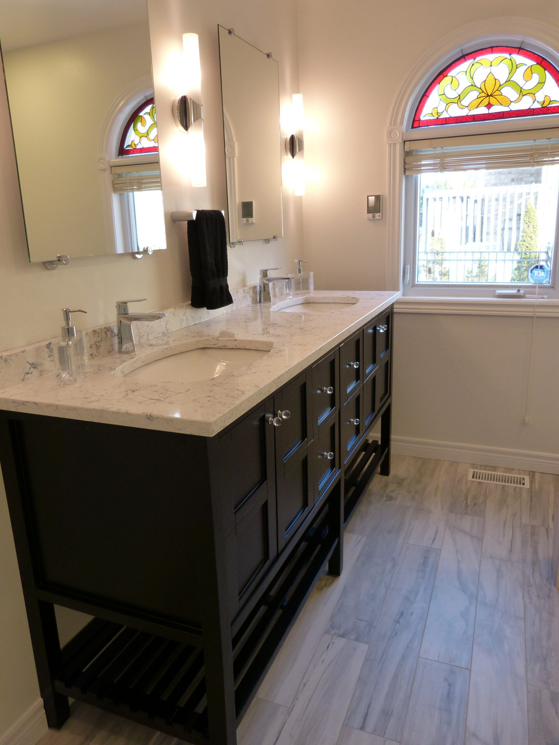 Bathroom with dark vanity, double sinks, large mirror, and arched window with stained glass.