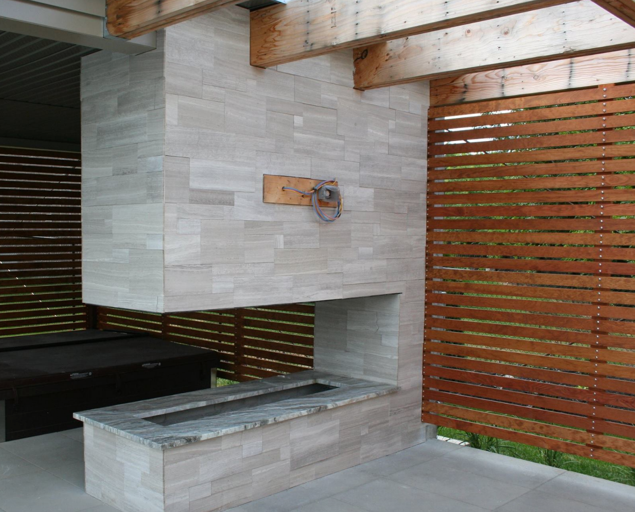 Outdoor fireplace with stone facade, under a wooden canopy, with a horizontal slatted wood fence.