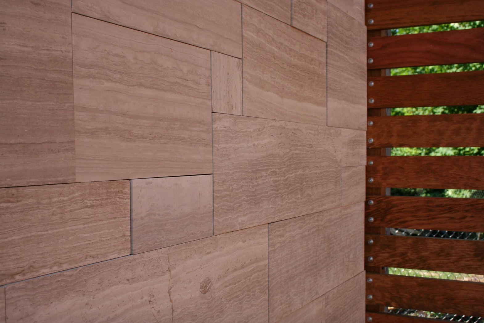 Brown sandstone wall with horizontal grain, adjacent to a wooden slat fence with glimpses of green foliage.