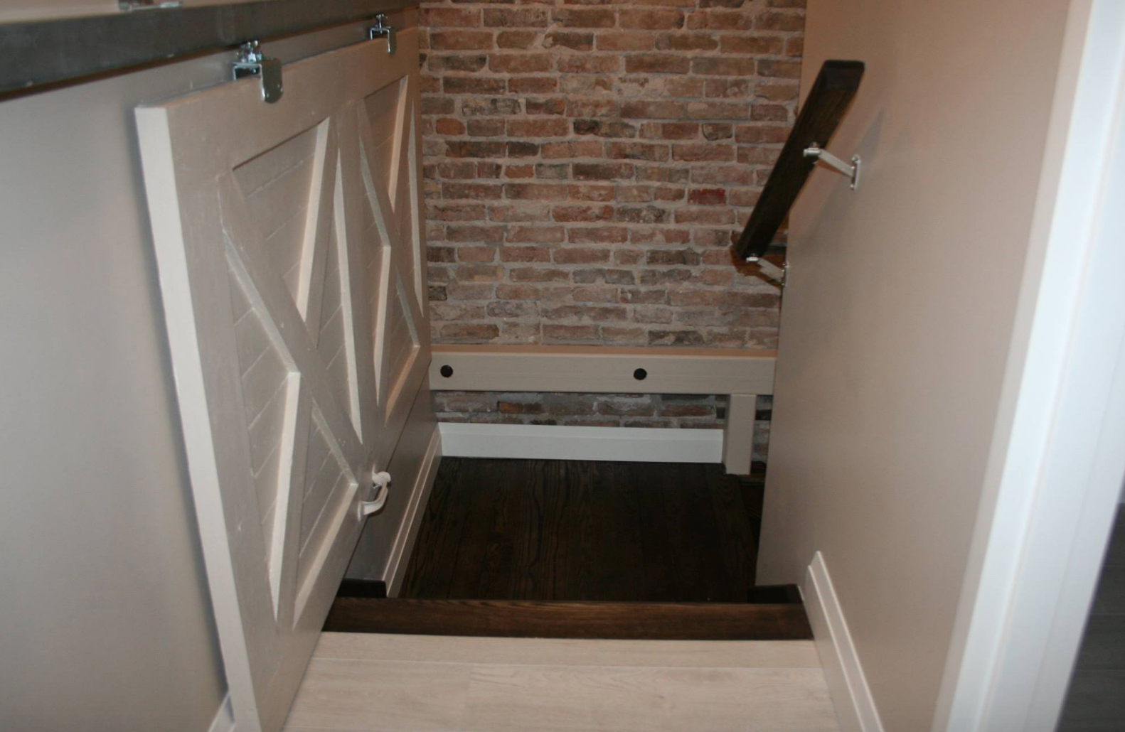 White barn door opens to reveal basement stairs with brick wall. Handrail on right.