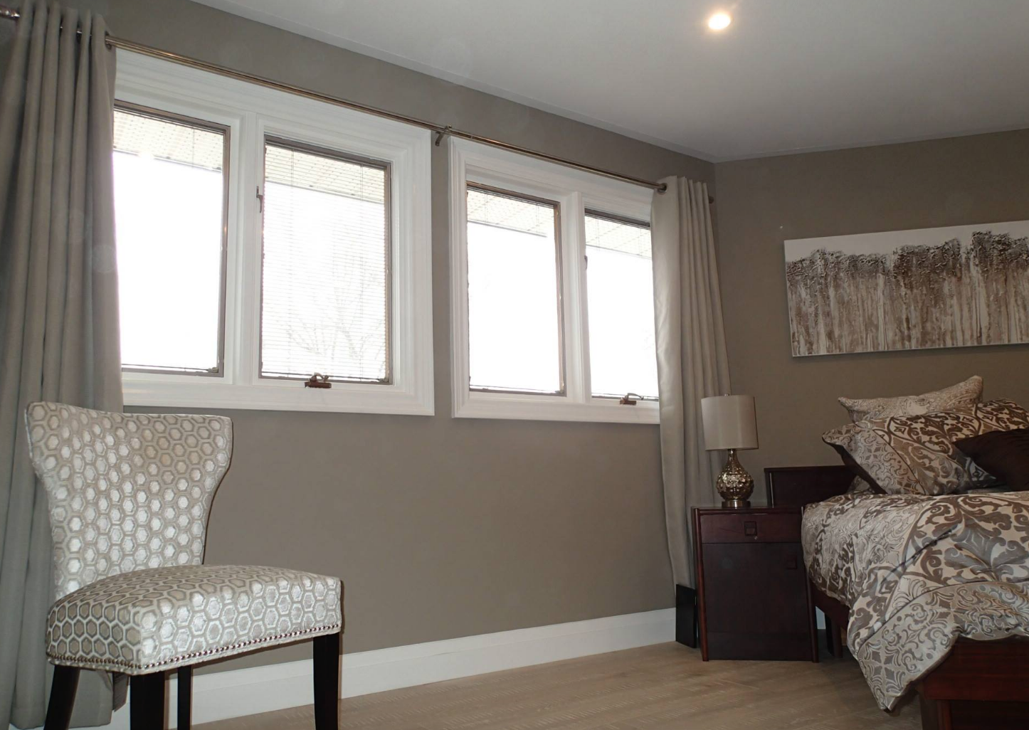 Bedroom with gray walls, three windows with light curtains, bed, chair, and artwork.