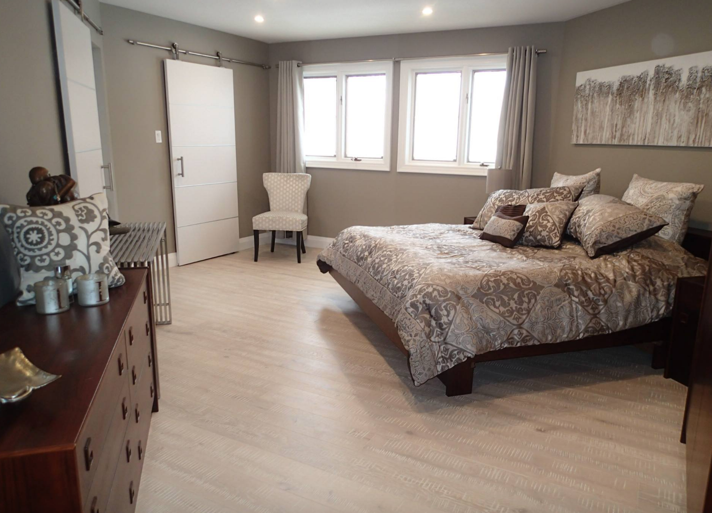 Bedroom with a bed, dresser, chair, and sliding door. Light wood floors, gray walls, and windows.