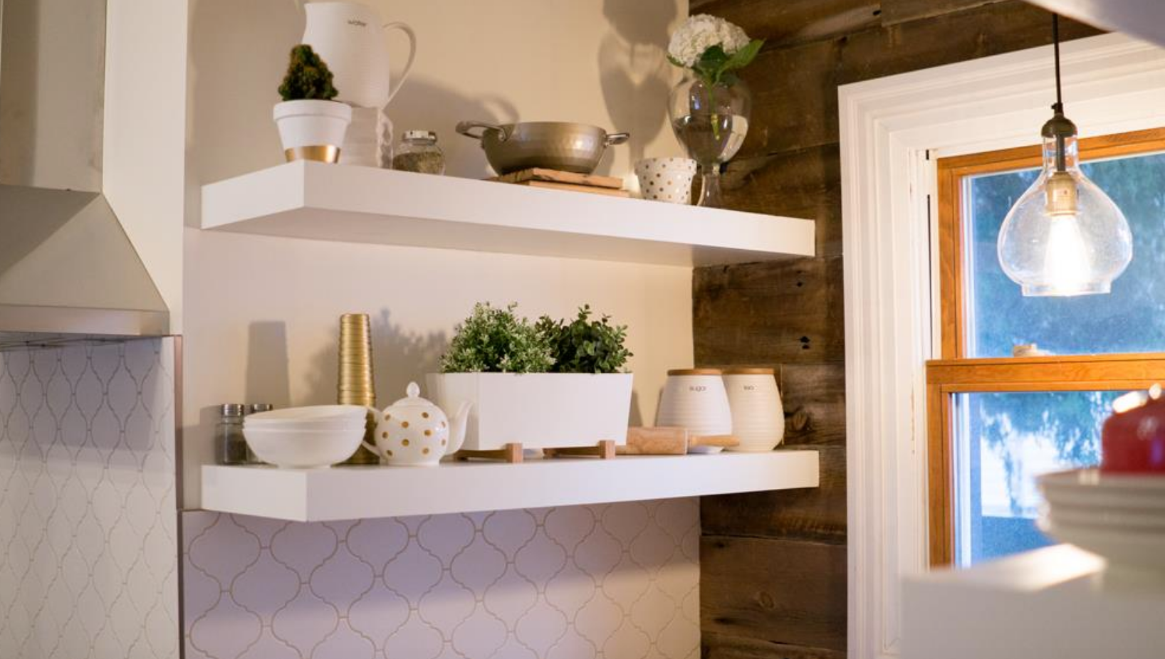 White floating shelves in a kitchen, stocked with decorative items. A light fixture hangs by a window.