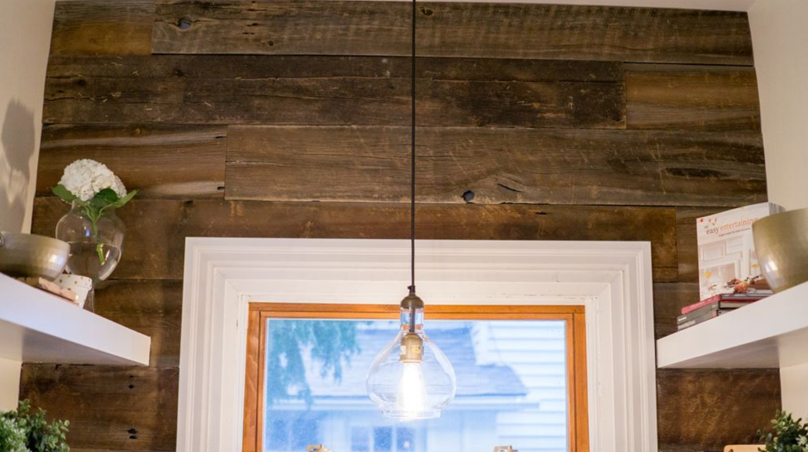 Wooden accent wall above a window, a light fixture hangs down. Shelves with decor on either side.