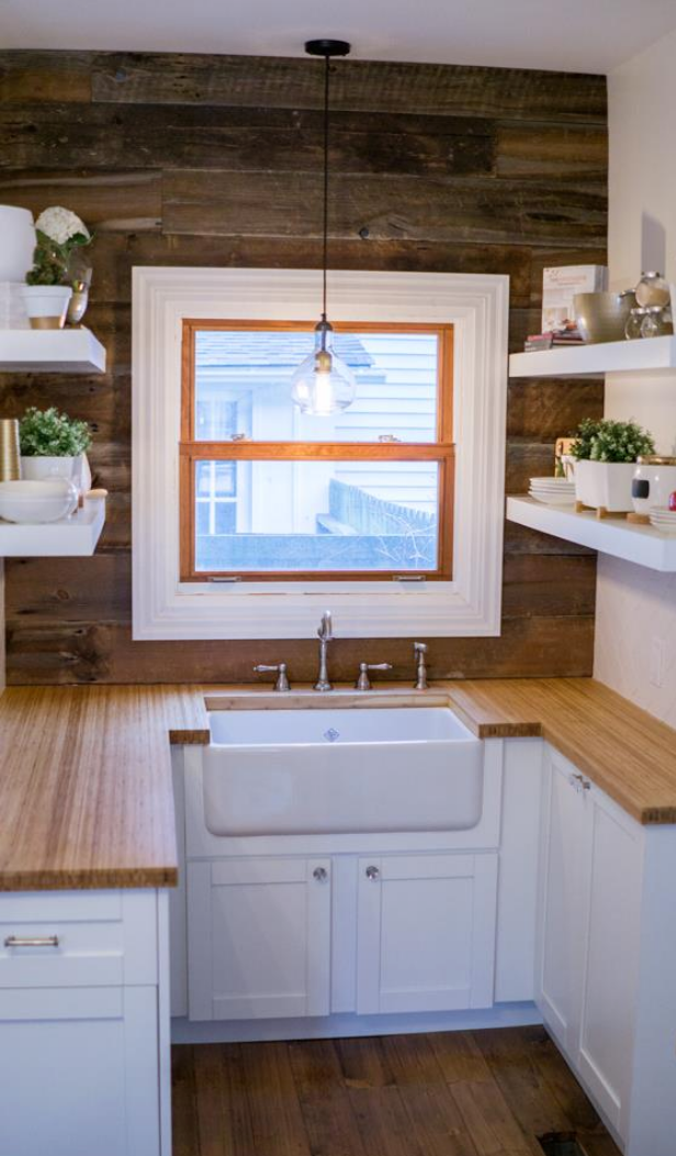 Kitchen with farmhouse sink, wooden counters, and shelves; a wooden accent wall, window, and hanging light.