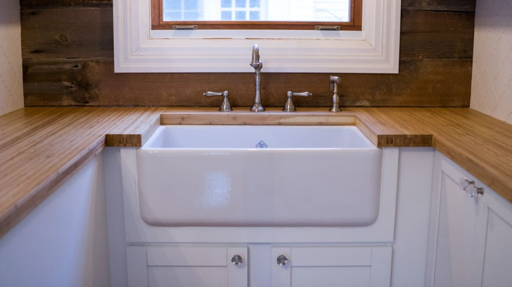 White farmhouse sink with wooden countertop in a small kitchen, under a window.