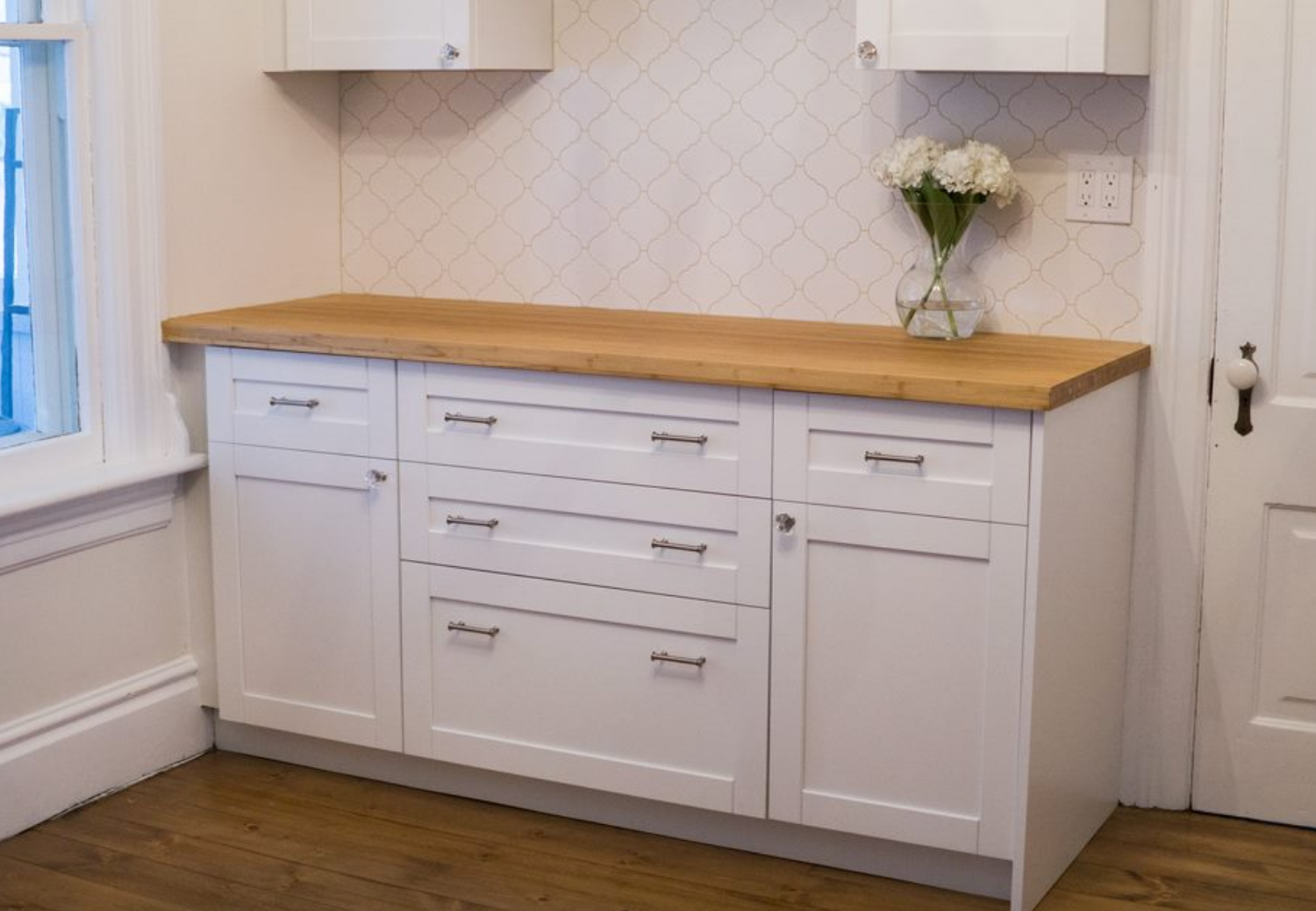 White cabinet with wood countertop and diamond tile backsplash; flowers on countertop.